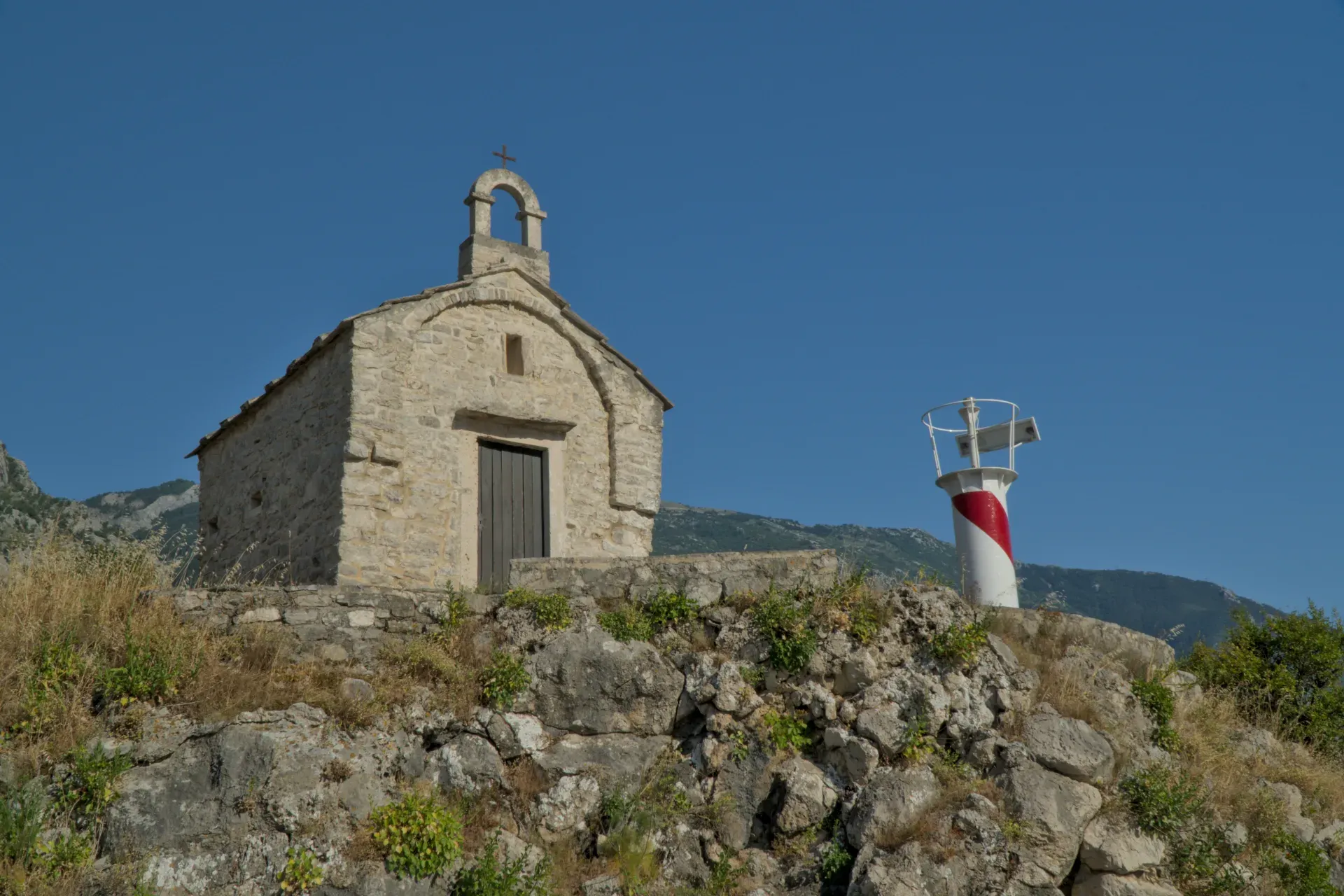 The stone Church of St. Elias in Dobrota, Montenegro, with a red and white lighthouse under a blue sky.