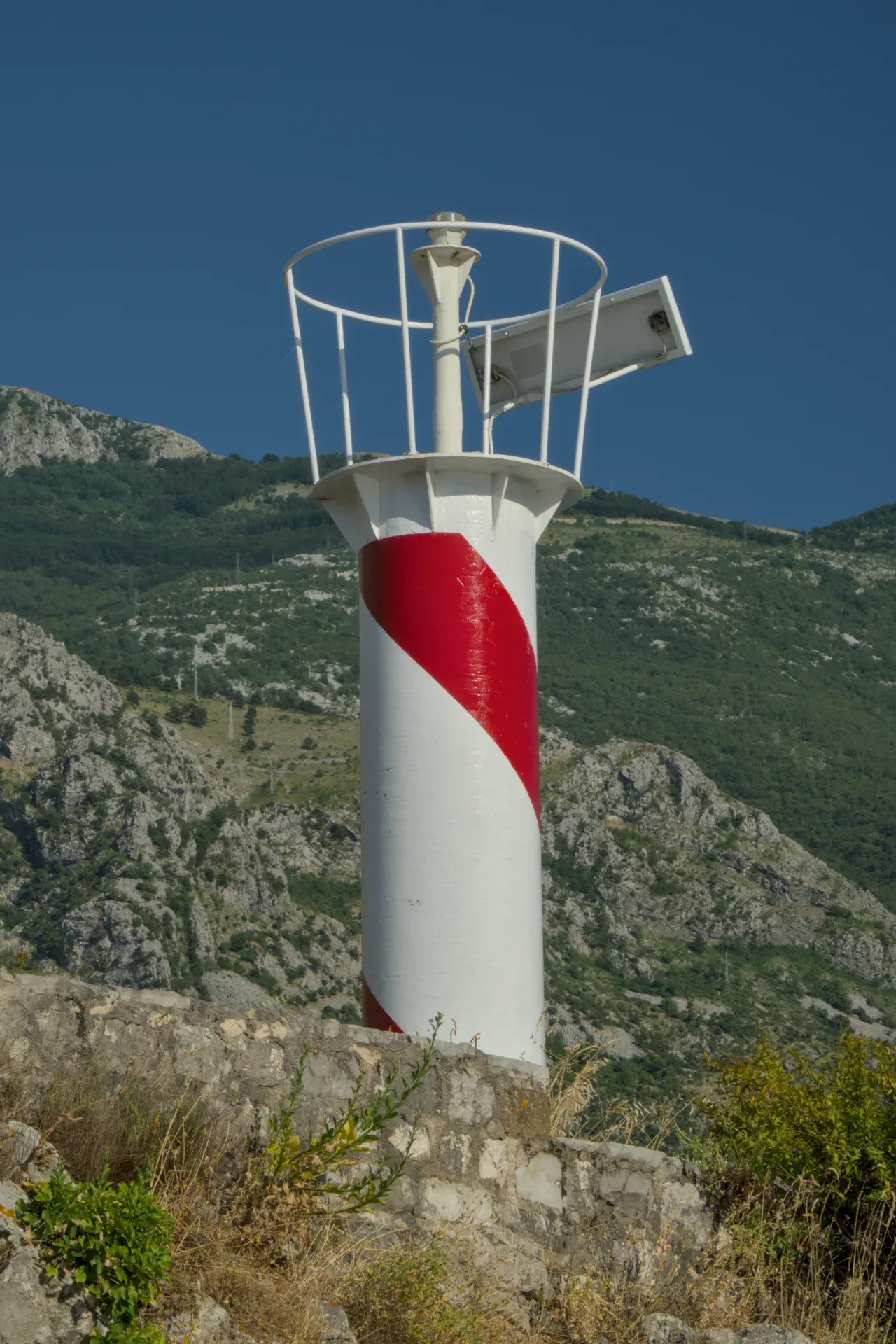 Un llamativo faro rojo y blanco se alza sobre una roca contra un cielo azul con montañas verdes al fondo.