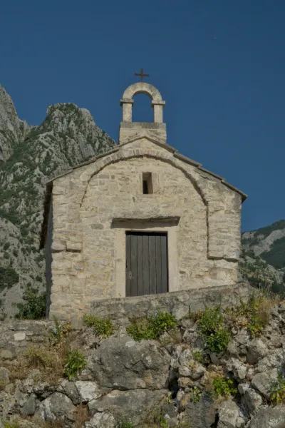 The Church of Saint Elias, a stone building with a wooden door, stands on a rocky slope under a blue sky in Dobrota, Montenegro.