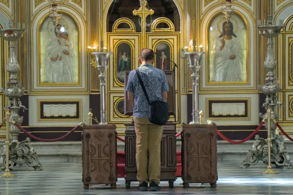 A man stands devoutly before an Orthodox iconostasis adorned with golden embellishments and icons.