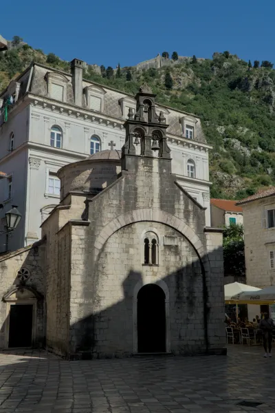 An old stone church with a bell tower and arched facade stands before a mountain fortress in Kotor.