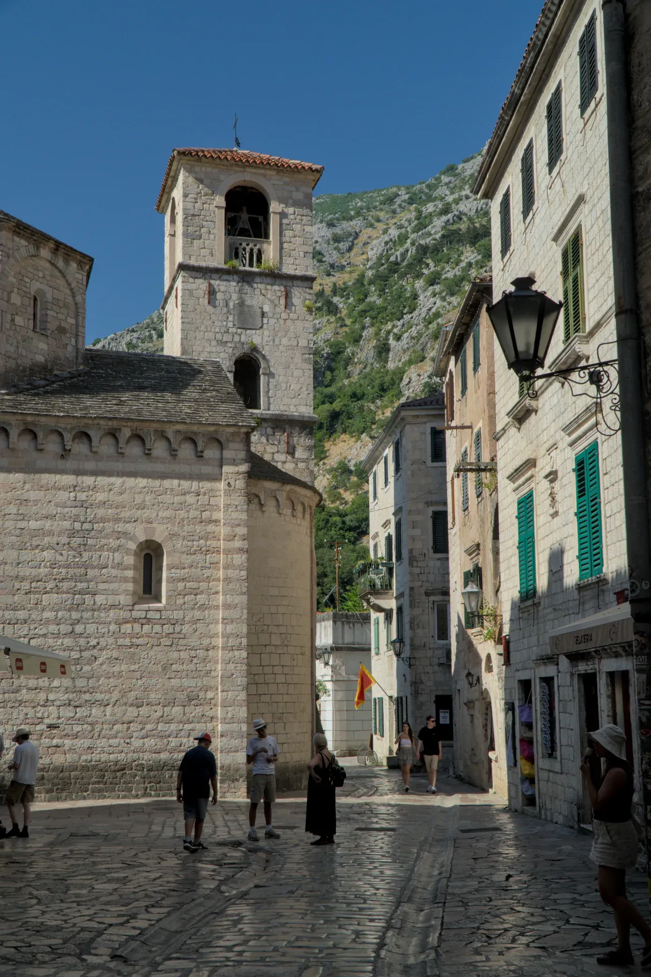 A narrow, cobblestone alley in the old town of Kotor, Montenegro, with historic stone buildings and a church tower in the background.