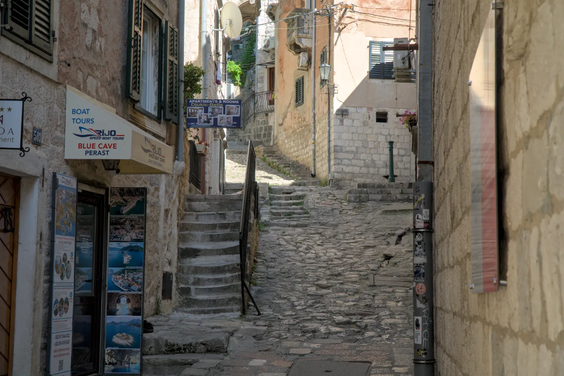 A steep, cobblestone alley in Kotor, Montenegro, with stone buildings and a stone staircase leading upwards.
