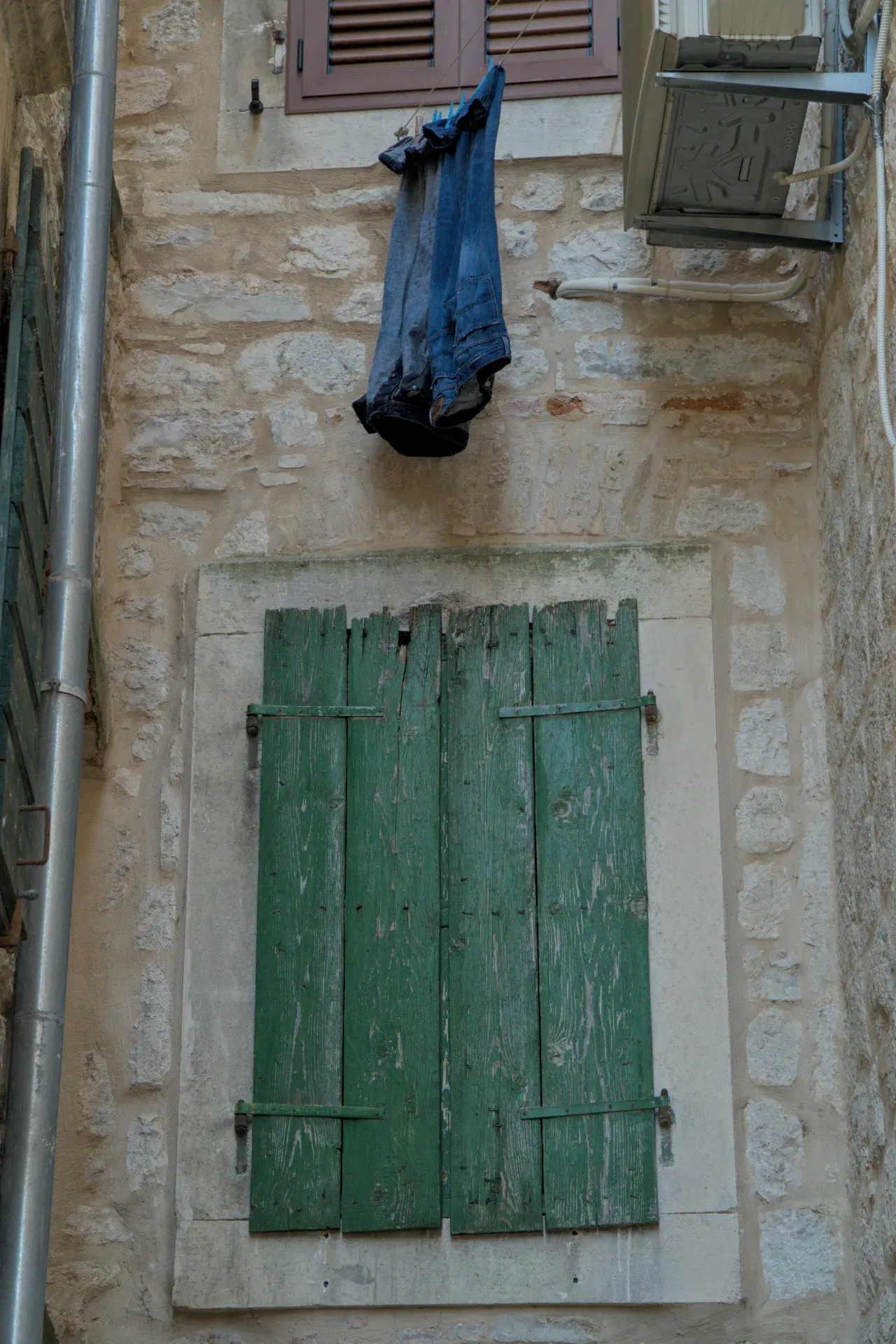A window with green wooden shutters and jeans hanging to dry on a rustic stone wall in Kotor.