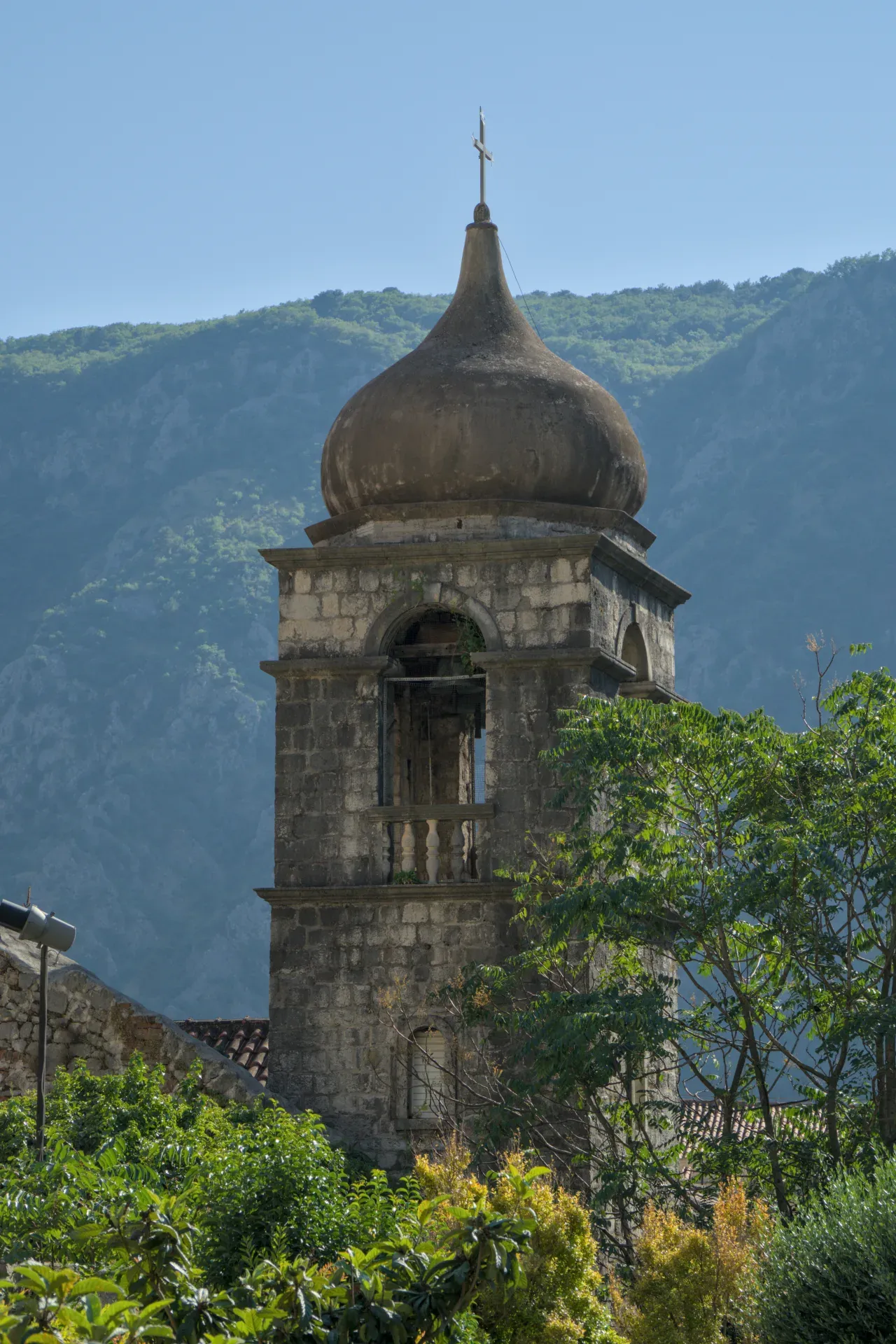 Histórica torre de iglesia de piedra con cúpula y cruz, frente a montañas boscosas y cielo azul en Kotor.