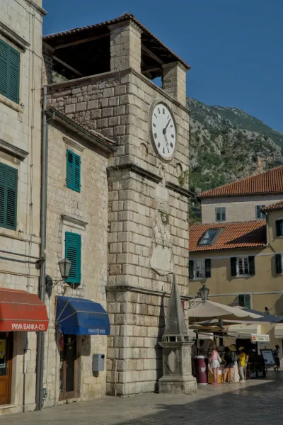 A stone clock tower in Kotor, Montenegro, with green shutters and a mountain in the background.