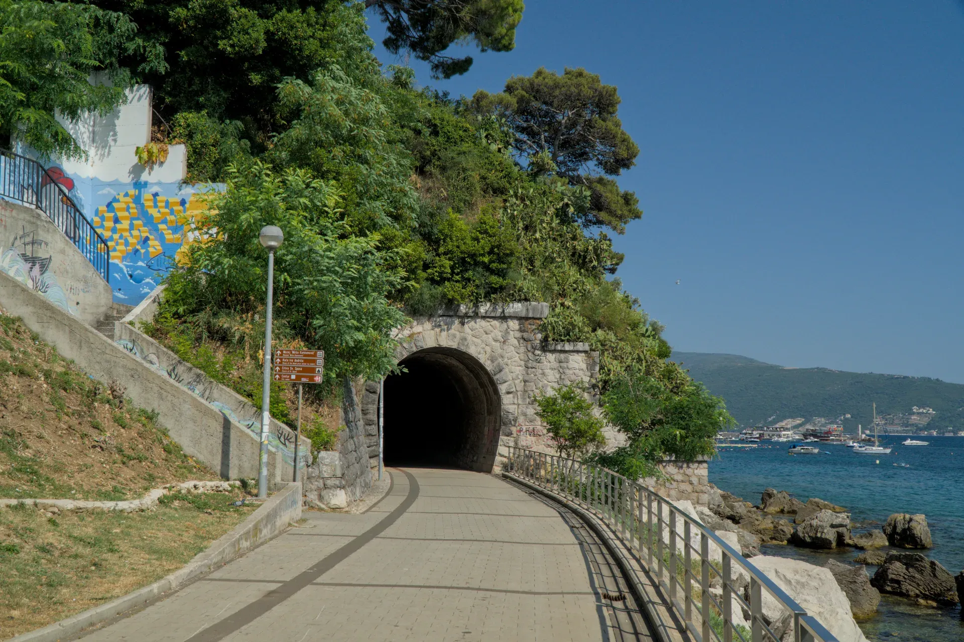 Ein Steintunnel mit beleuchtetem Eingang führt entlang der Küste von Herceg Novi, Montenegro, mit Blick auf das Meer und die Berge.