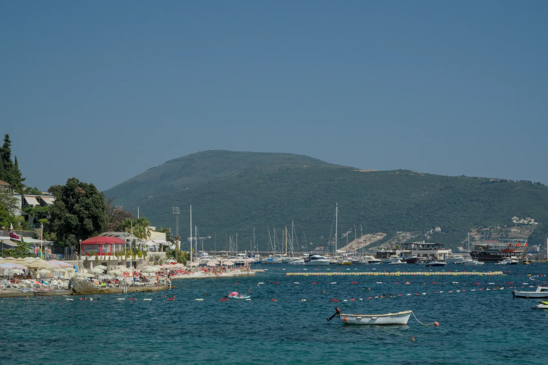 Boats in Herceg Novi harbor, Montenegro, with a bustling beach promenade and green hills in the background.