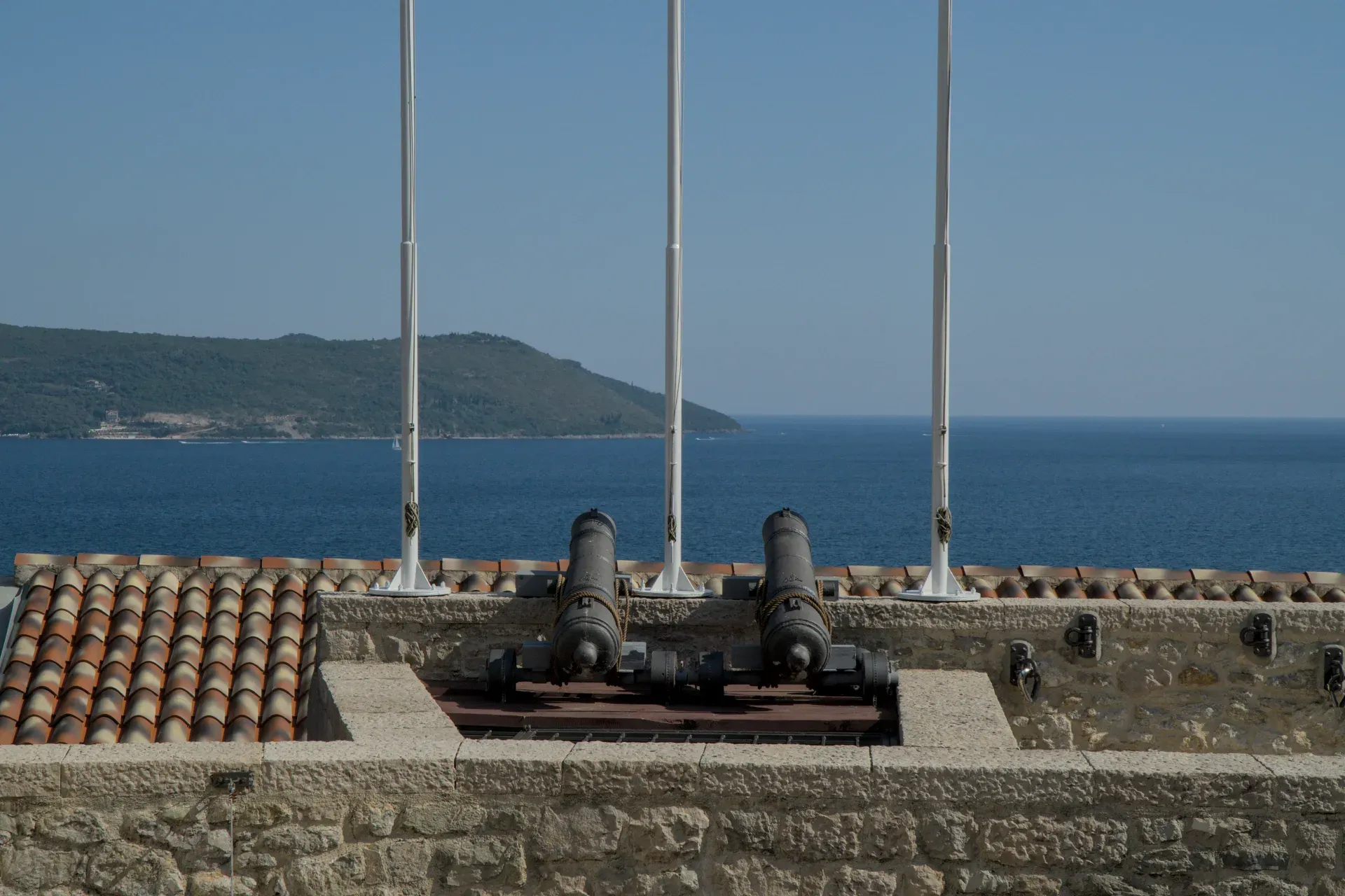Due vecchi cannoni si ergono su un muro di fortezza in pietra che domina il mare blu e la costa verde sotto un cielo limpido.