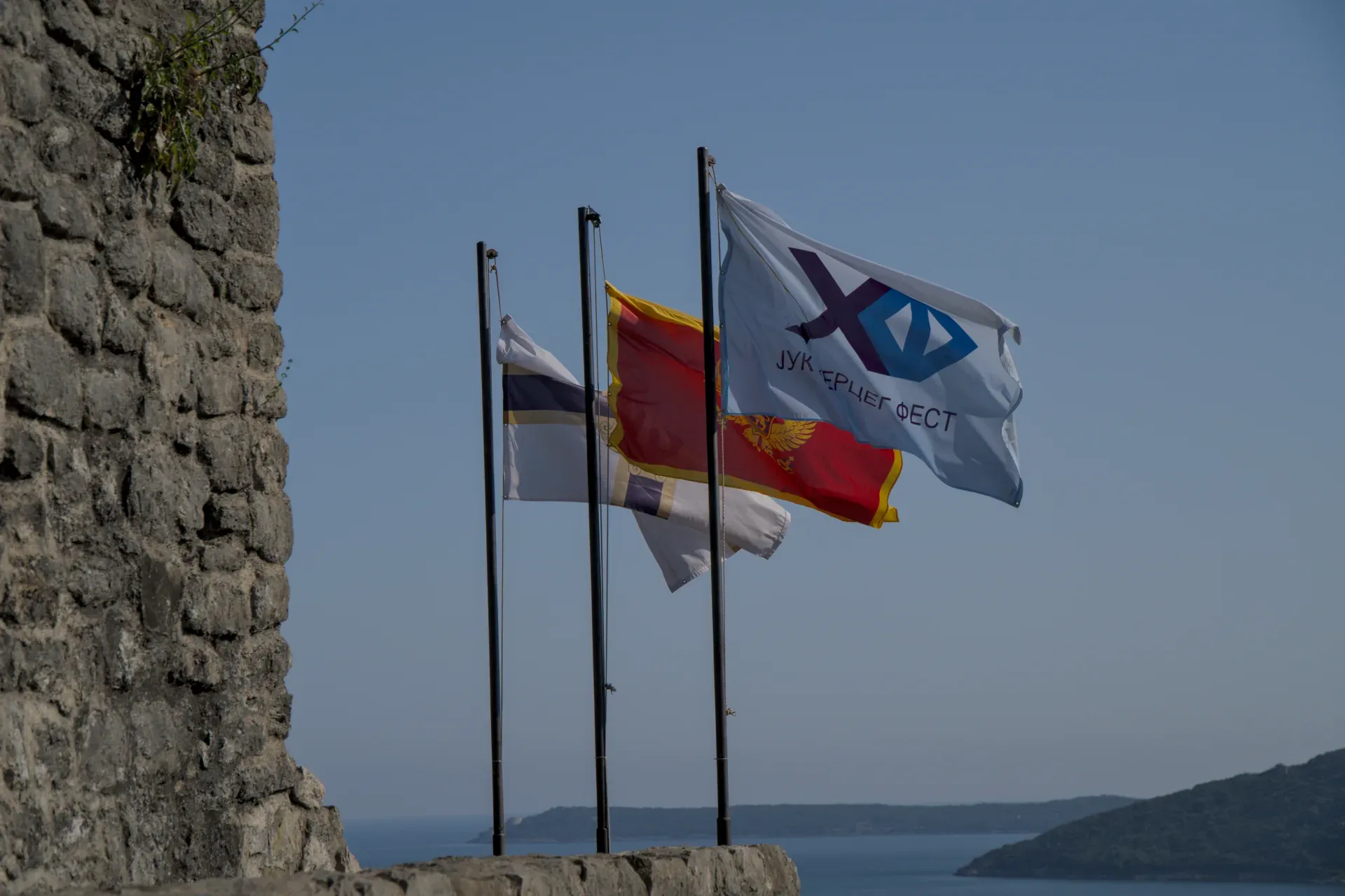 Three flags wave in the wind over the sea in Herceg Novi, next to an old stone wall.