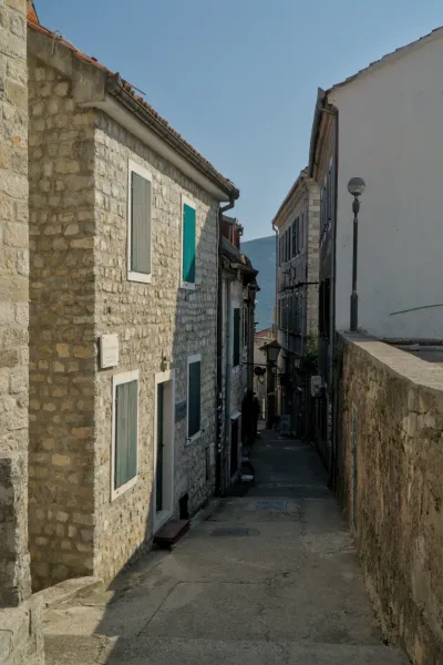 A narrow, cobblestone alley in Herceg Novi with old stone houses and green shutters, in the sunlight.