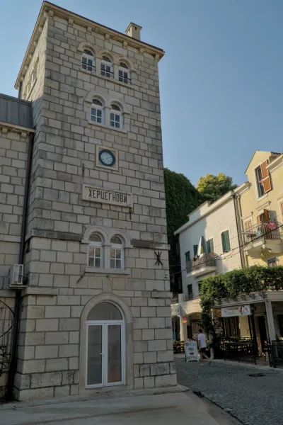A stone clock tower in Herceg Novi, Montenegro, featuring a blue clock face and Cyrillic inscription above an archway.