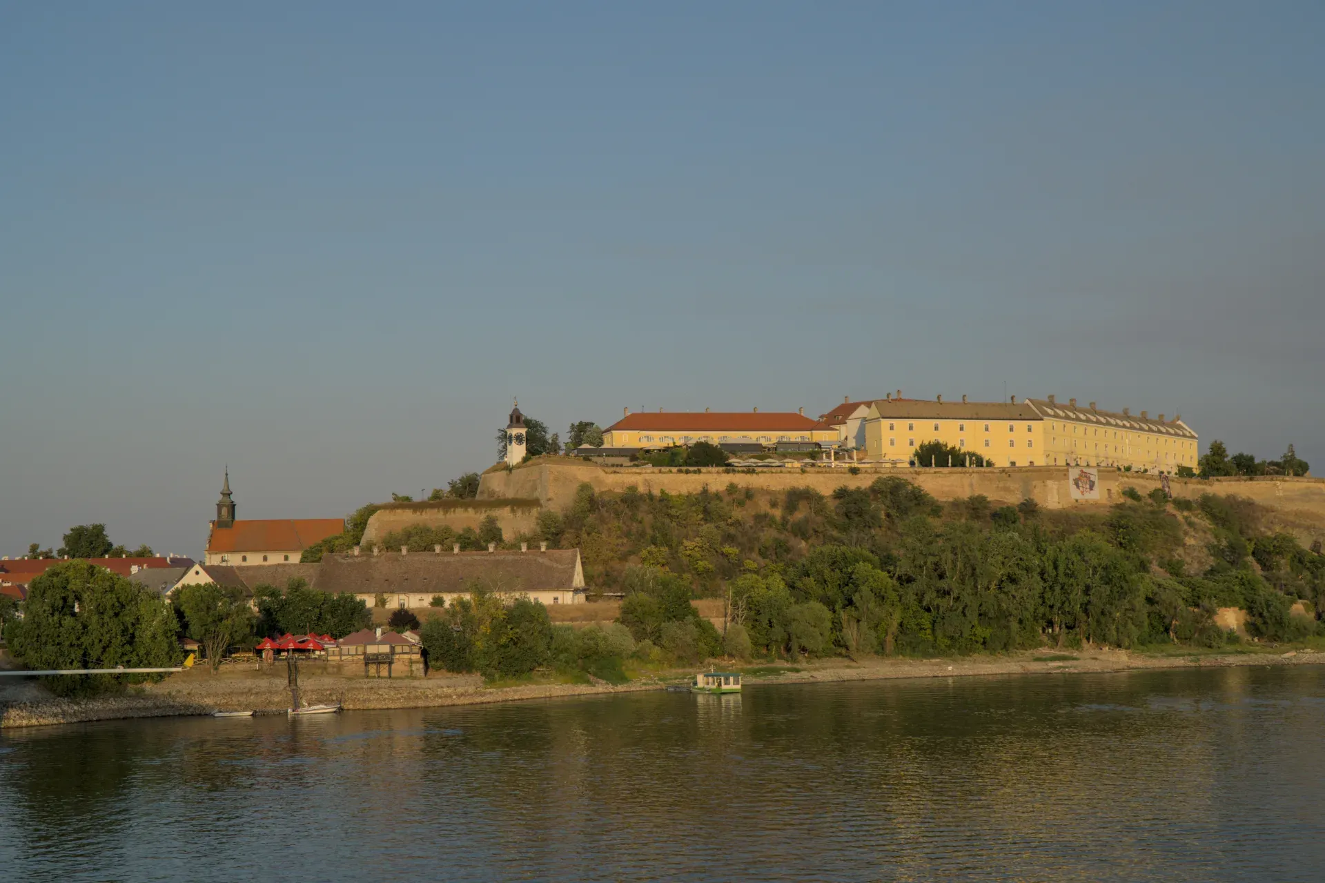 Petrovaradin Fortress, Serbia, in warm evening light, with river and trees in the foreground.