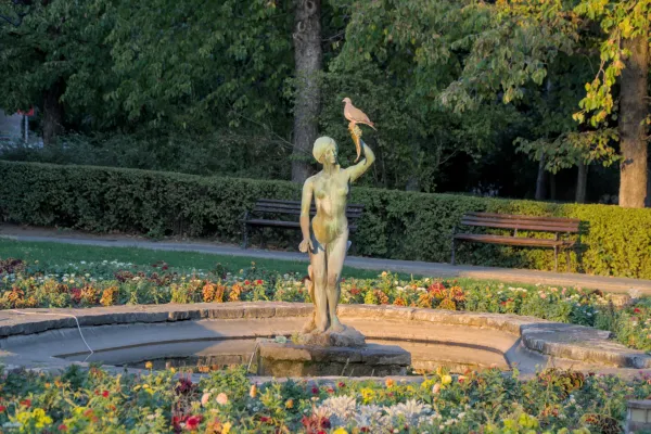Bronze statue of a girl with a cornucopia and a dove in Danube Park Novi Sad, Serbia, with flowers and trees in the background.