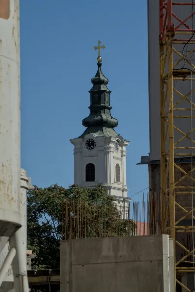 The bell tower of the Dormition Cathedral in Novi Sad rises against a clear blue sky, with its green roof and golden cross.