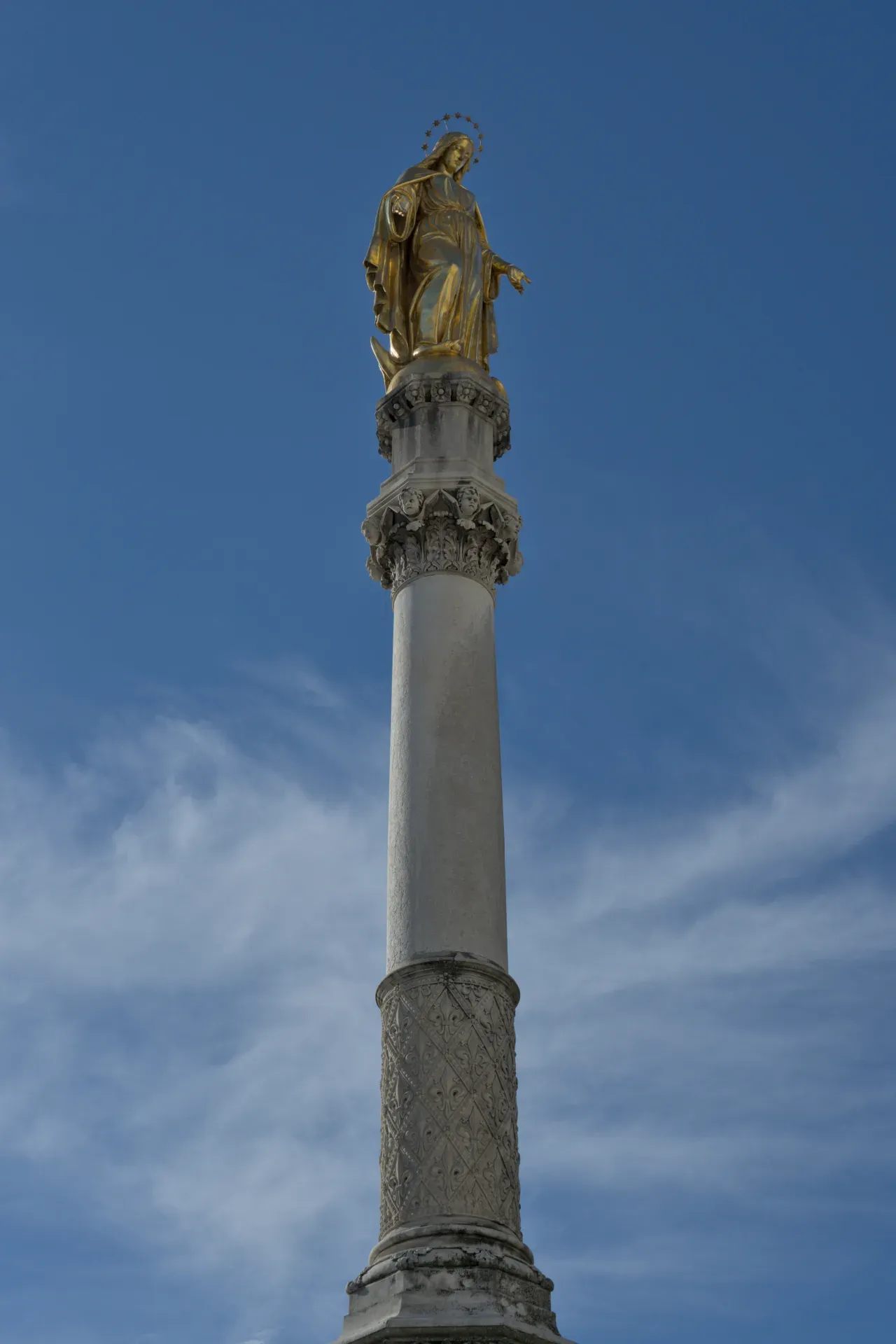 Statua dorata della Vergine Maria su una colonna finemente decorata, contro un cielo blu con nuvole sottili.