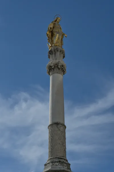 Goldene Marienstatue auf einer kunstvoll verzierten Säule vor einem blauen Himmel mit zarten Wolken.