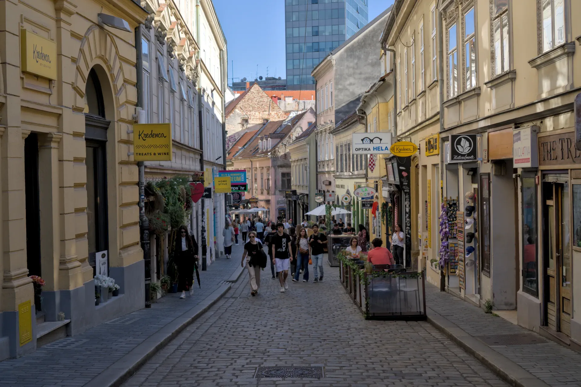 A sun-drenched, cobblestone alley in Zagreb, lined with shops and cafes, with people strolling by.