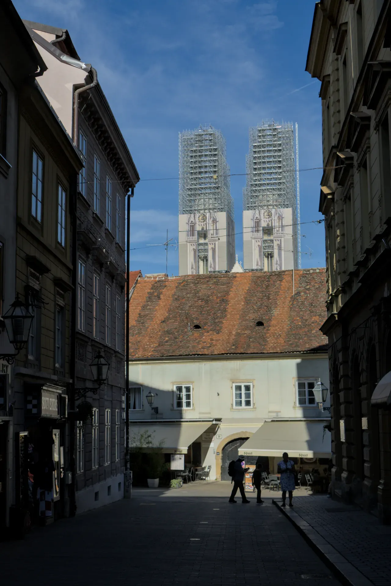 Architectural photograph of Zagreb Cathedral standing in scaffolding against a blue sky, with people walking through an alley.