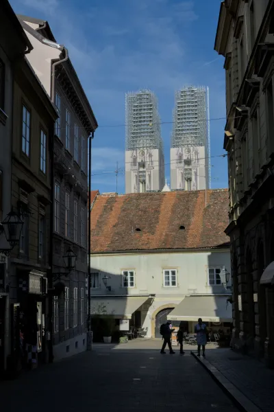 Architekturfotografie der Kathedrale von Zagreb, die im Gerüst vor blauem Himmel steht, mit Menschen, die durch eine Gasse gehen.