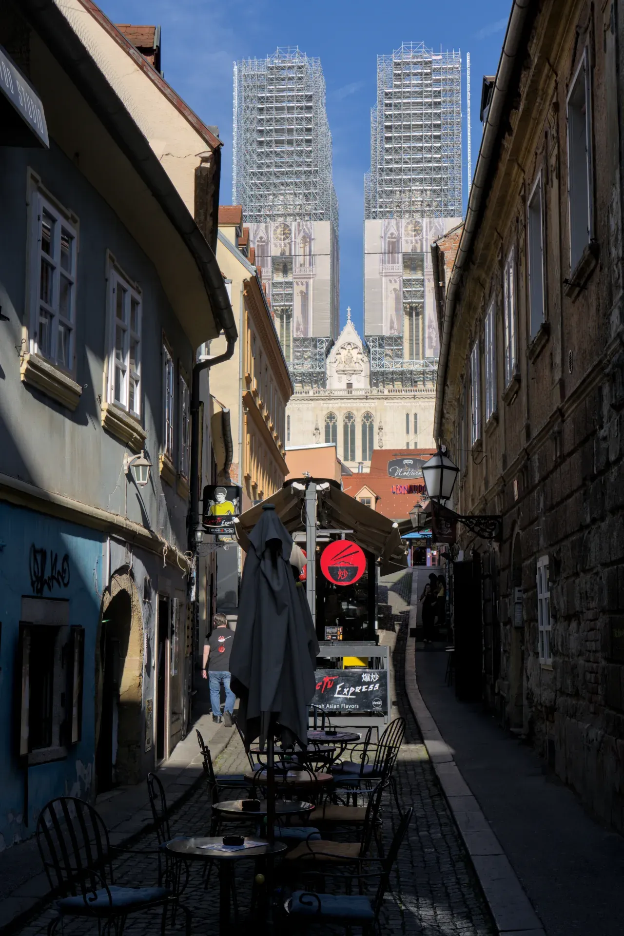 Impresivna Zagrebačka katedrala, delimično zaklonjena građevinskim skelama, uzdiže se iznad šarmantnog uskog prolaza sa stolovima u kafićima i fasadama u prvom planu.