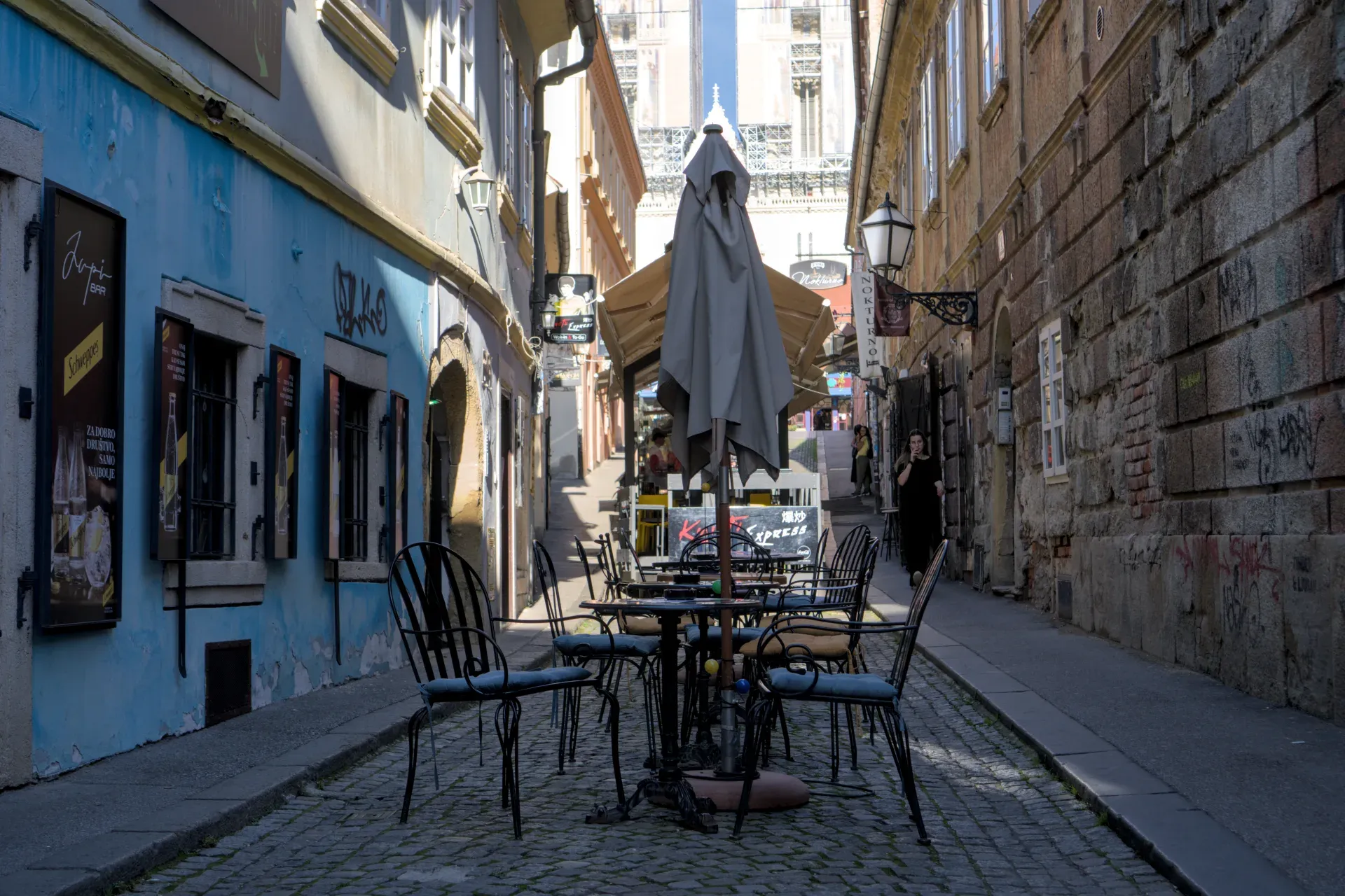 A narrow alley in Zagreb, lined with buildings with blue and stone facades, with cafe tables in the foreground.