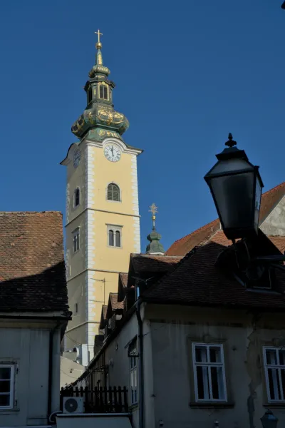 Ein Porträtfoto eines gelben Kirchturms mit grüner Kuppel und goldenen Verzierungen, eingefangen im klaren blauen Himmel über Zagreb.