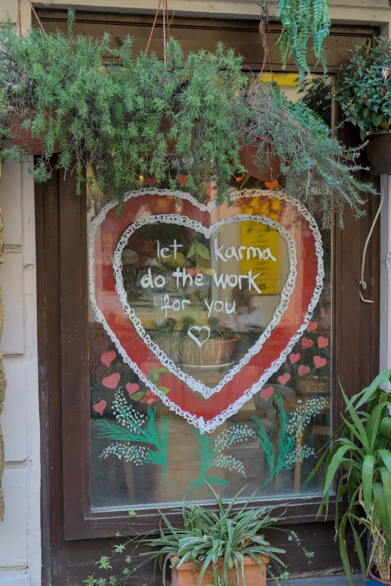 A hand-painted heart with the inscription 'Let karma do the work for you' on a window, surrounded by plants and heart-shaped accents.