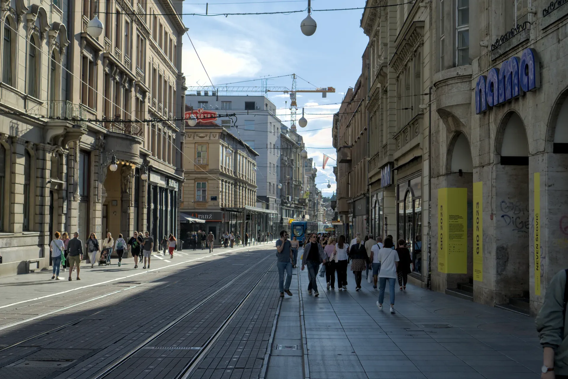 Eine sonnige Straße in Zagreb mit Straßenbahnschienen und historischen Gebäuden, belebt von Spaziergängern.