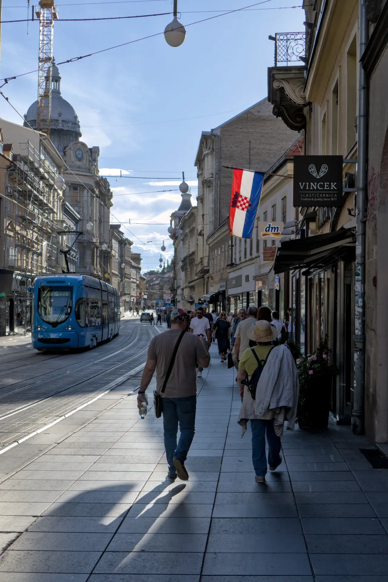 Eine blaue Straßenbahn fährt auf den Gleisen durch eine Straße in Zagreb, flankiert von Gebäuden und Fußgängern.