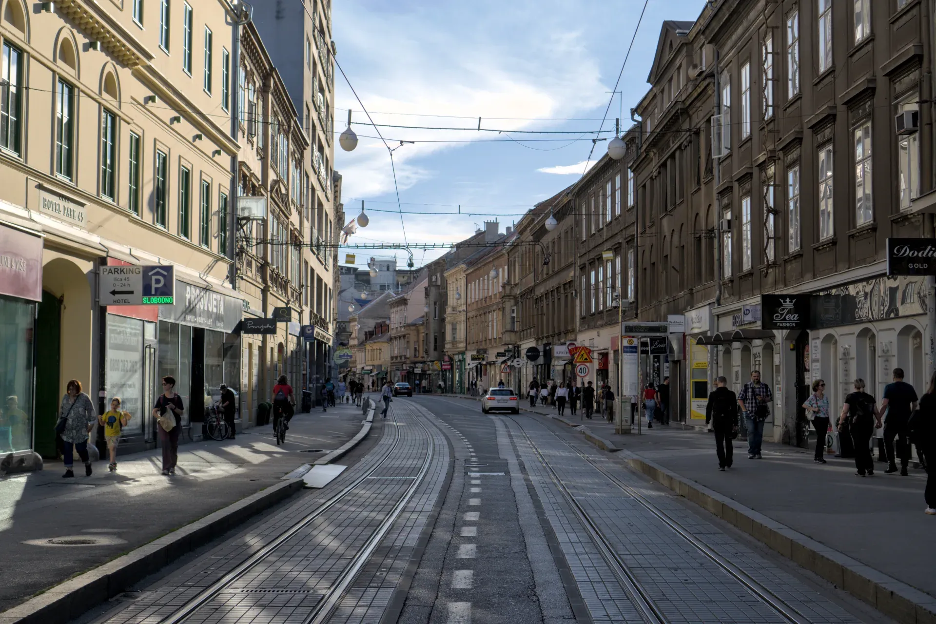 Eine urbane Straße in Zagreb mit historischen Gebäuden, Straßenbahnschienen und Menschen, die an einem sonnigen Tag spazieren gehen.