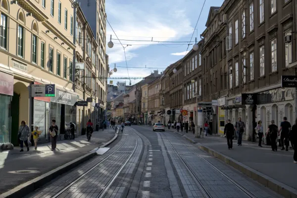 Eine urbane Straße in Zagreb mit historischen Gebäuden, Straßenbahnschienen und Menschen, die an einem sonnigen Tag spazieren gehen.