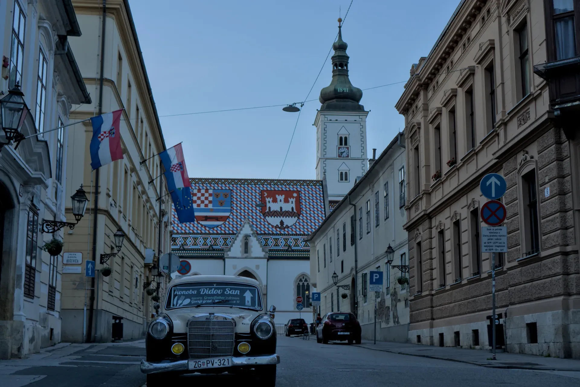 Klasični automobil i crkva Svetog Marka u Zagrebu sa hrvatskim zastavama u večernjem svetlu.