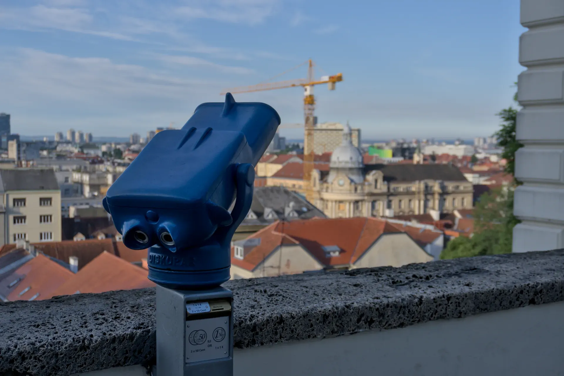 Plavi dvogled za posmatranje nadvisuje slikoviti horizont Zagreba sa crvenim krovovima i dizalicom u pozadini.