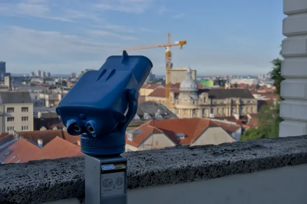 Ein blaues Beobachtungsfernglas thront vor der malerischen Skyline von Zagreb mit roten Dächern und einem Kran im Hintergrund.