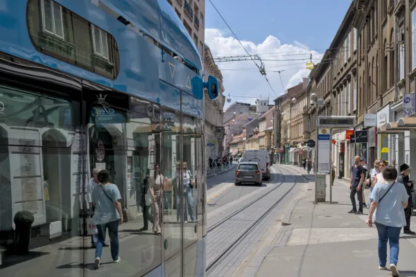 Eine blaue Straßenbahn fährt durch die belebte Straße von Zagreb, reflektiert die Gebäude und Menschen, mit historischer Architektur im Hintergrund.