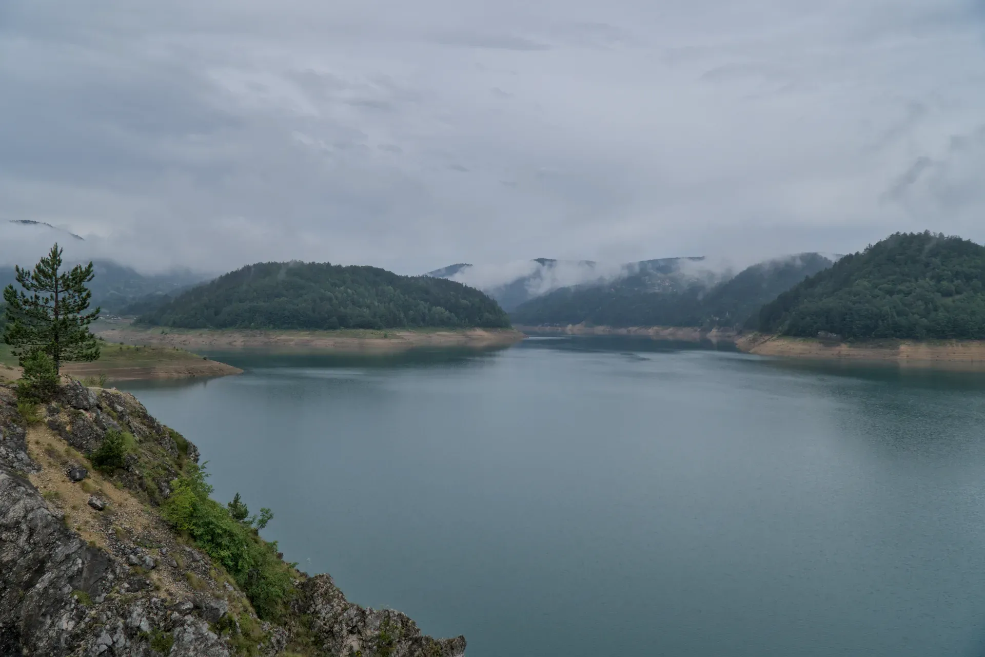 Dense clouds and mist over Zaovine Lake in Serbia, with green hills in the background.