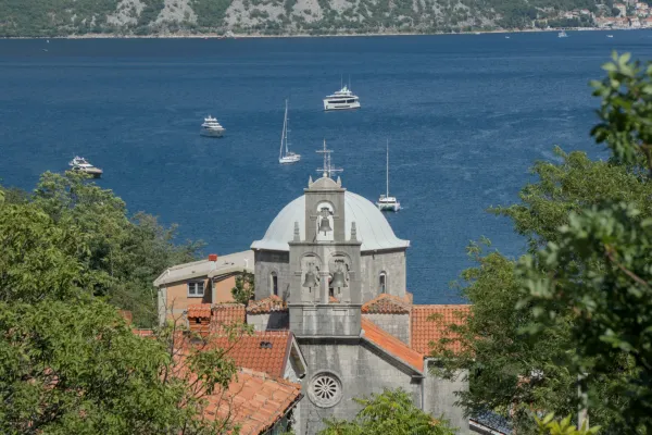 A church with a dome and bell tower stands before the Bay of Kotor, Montenegro, near Morinj, with yachts on the water.