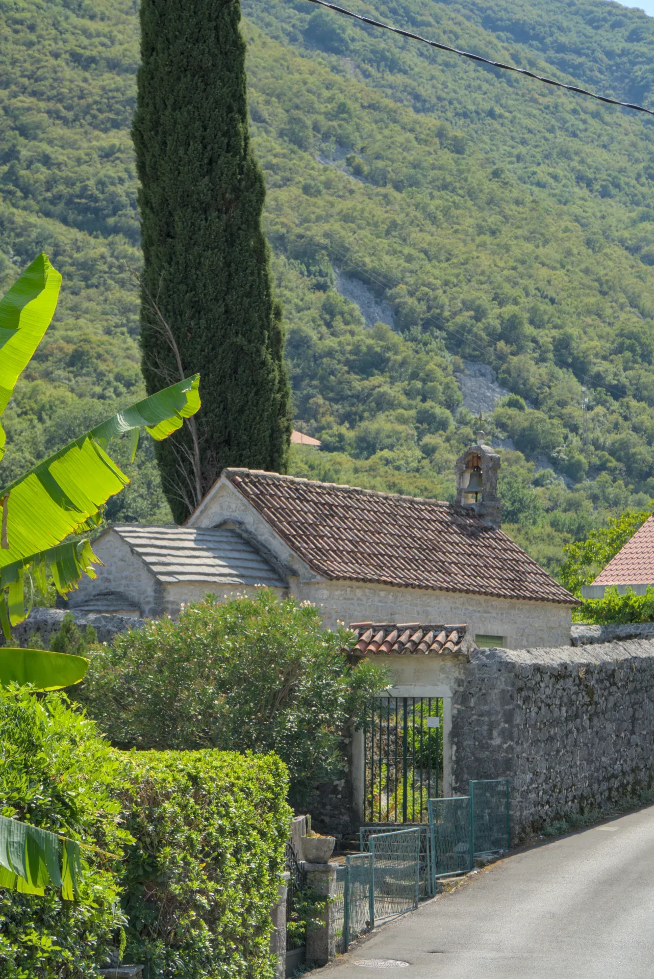 A small stone church with terracotta tiled roofs, flanked by lush greenery and a tall cypress tree, near Kotor, Montenegro.