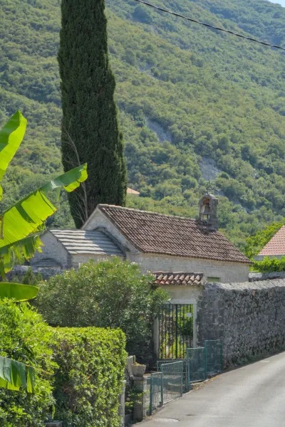 A small stone church with terracotta tiled roofs, flanked by lush greenery and a tall cypress tree, near Kotor, Montenegro.