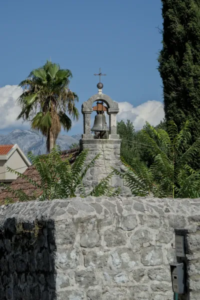 The bell tower of St. Trivun Catholic Church in Kotor, Montenegro, with palm trees and mountains in the distance.