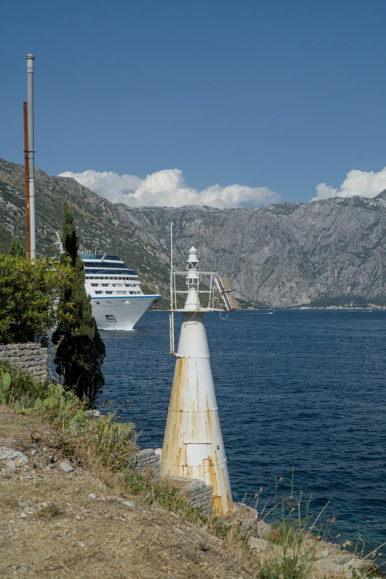 Izbledeli beli svetionik stoji na obali Kotorskog zaliva, dok veliki kruzer plovi vodom.