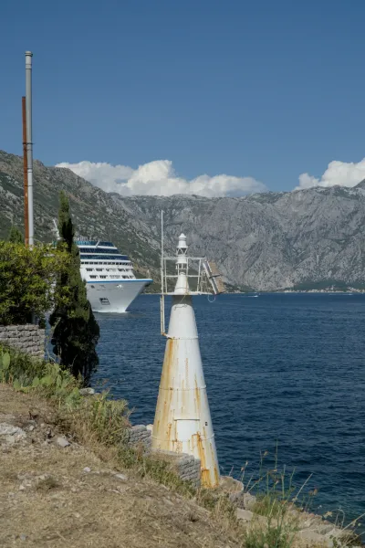 A weathered white lighthouse stands on the shore of Kotor Bay, with a large cruise ship sailing on the water.