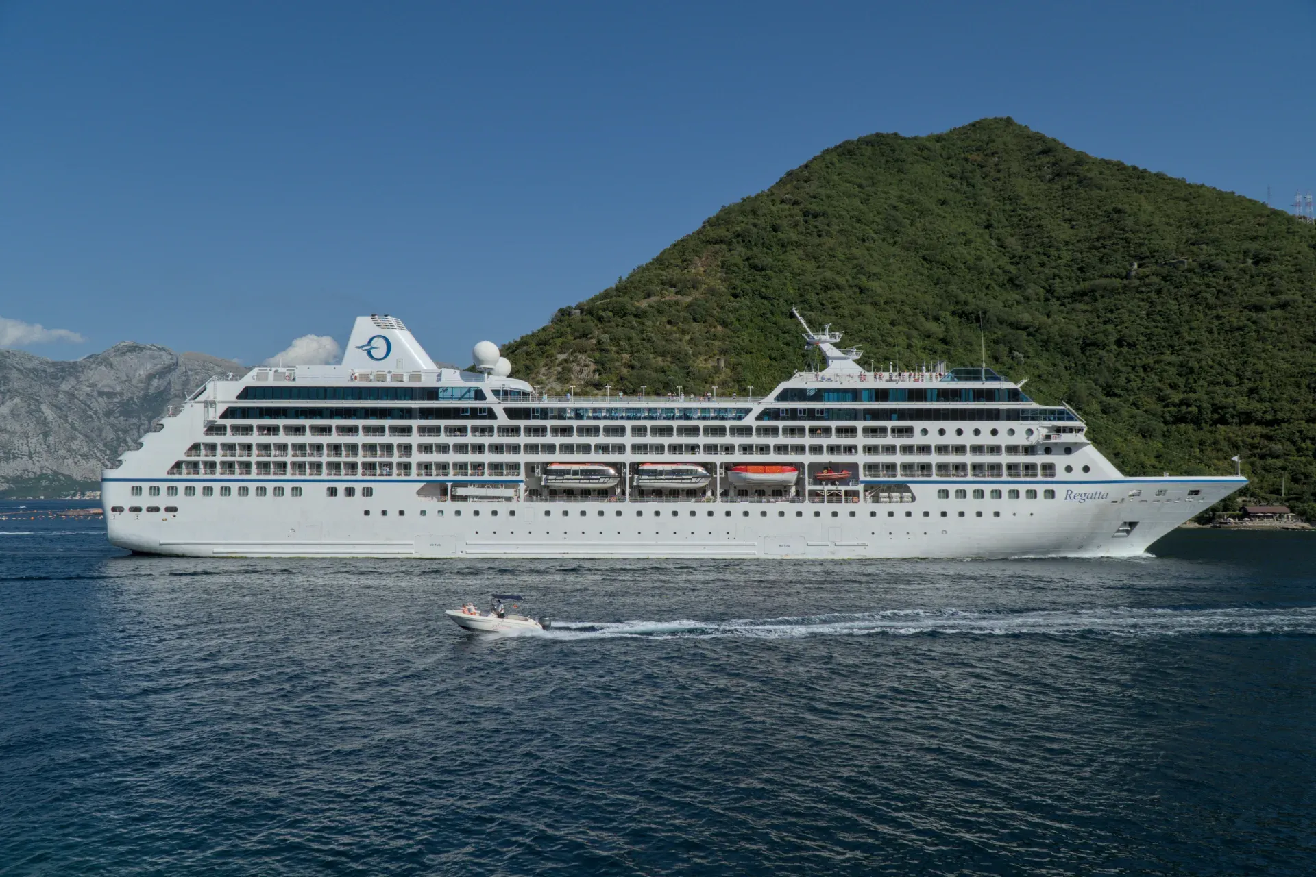 A large white cruise ship departs Kotor Bay as a small speedboat navigates the water.