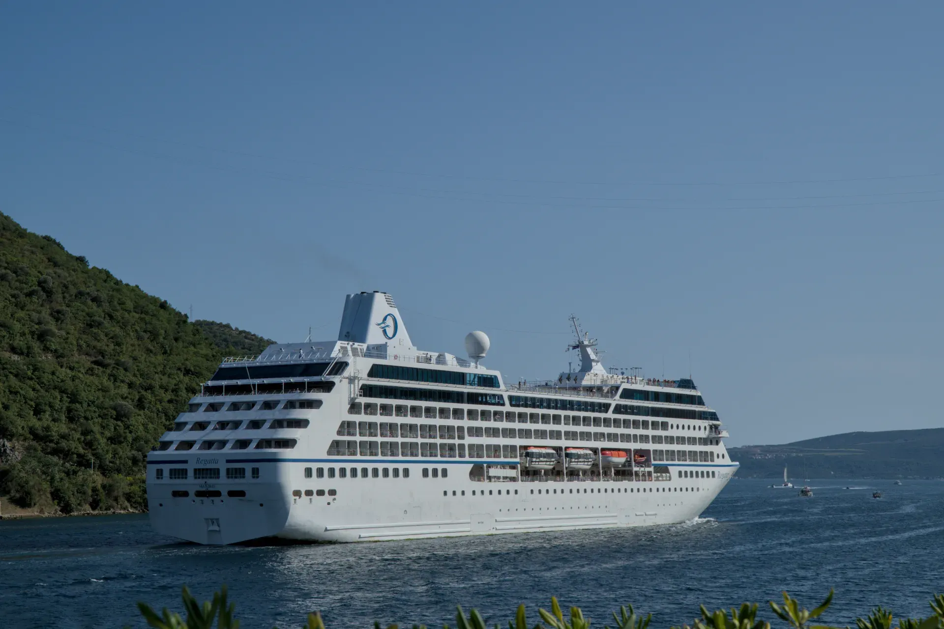 Una grande nave da crociera bianca che esce dalla Baia di Kotor sotto un cielo azzurro limpido, con le verdi colline del Montenegro sullo sfondo.