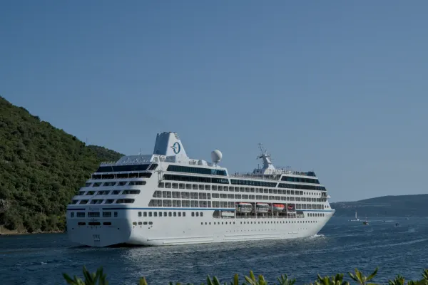 A large white cruise ship sailing out of Kotor Bay under a clear blue sky, with Montenegro's green hills in the background.