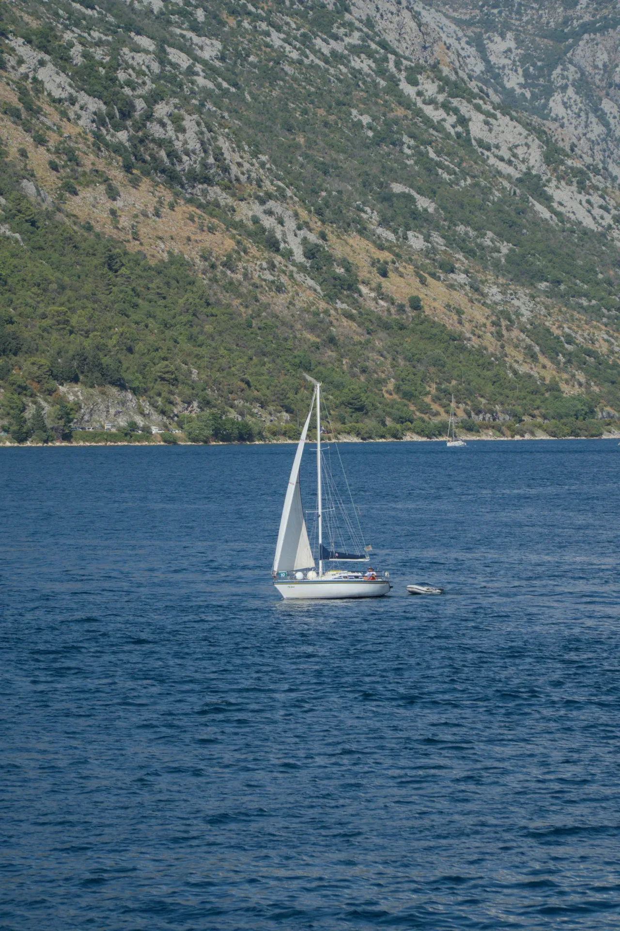 A small sailboat sails on the water off Kotor, Montenegro, with steep, vegetated mountains in the background.