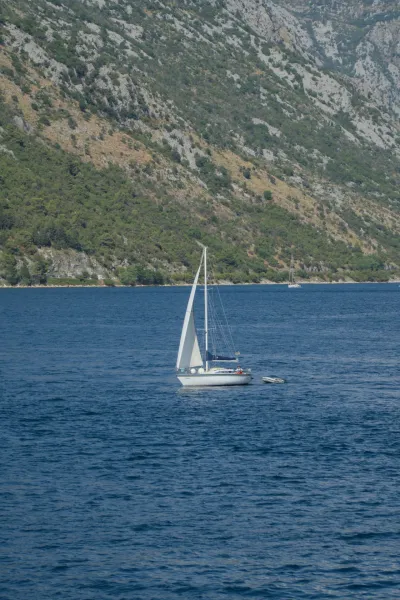 A small sailboat sails on the water off Kotor, Montenegro, with steep, vegetated mountains in the background.