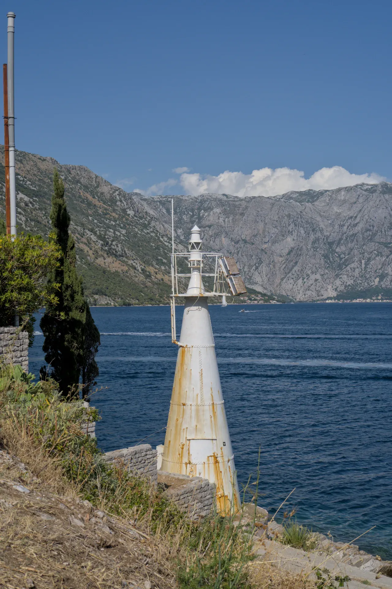 Faro blanco erosionado en la entrada de la Bahía de Kotor, Montenegro, con montañas y mar azul de fondo.