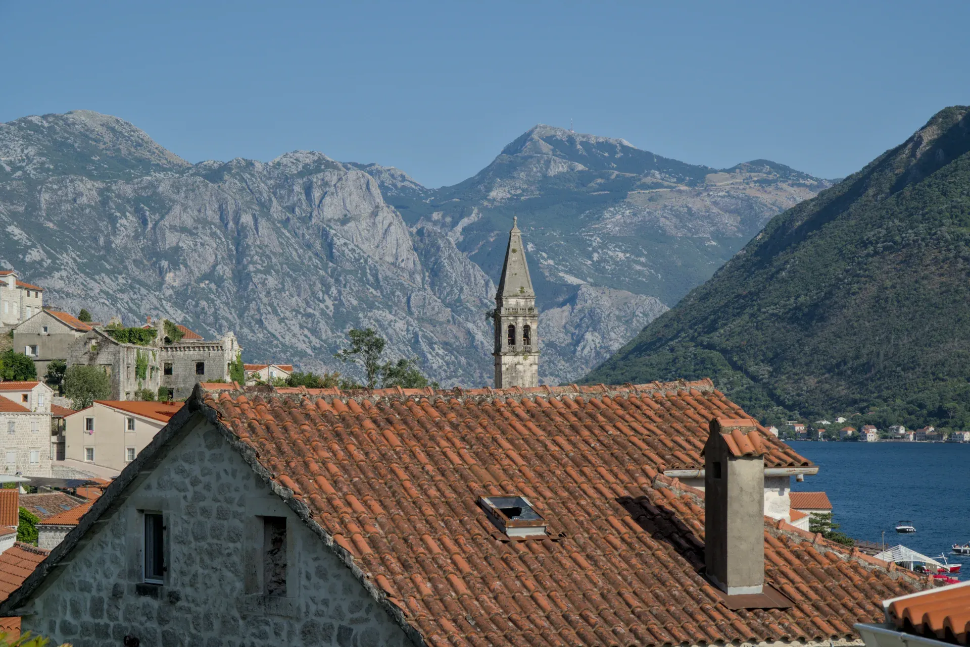 Perast, Montenegro: A view of the urban rooftops with a mountain backdrop, featuring a church tower and the water.